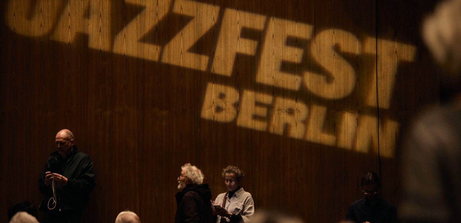 Der Schriftzug „Jazzfest Berlin“ wird an die Wand des Zuschauersaals des Haus der Berliner Festspiele projiziert. Zuschauer*innen stehen davor haben zum Teil ihre Plätze eingenommen.
