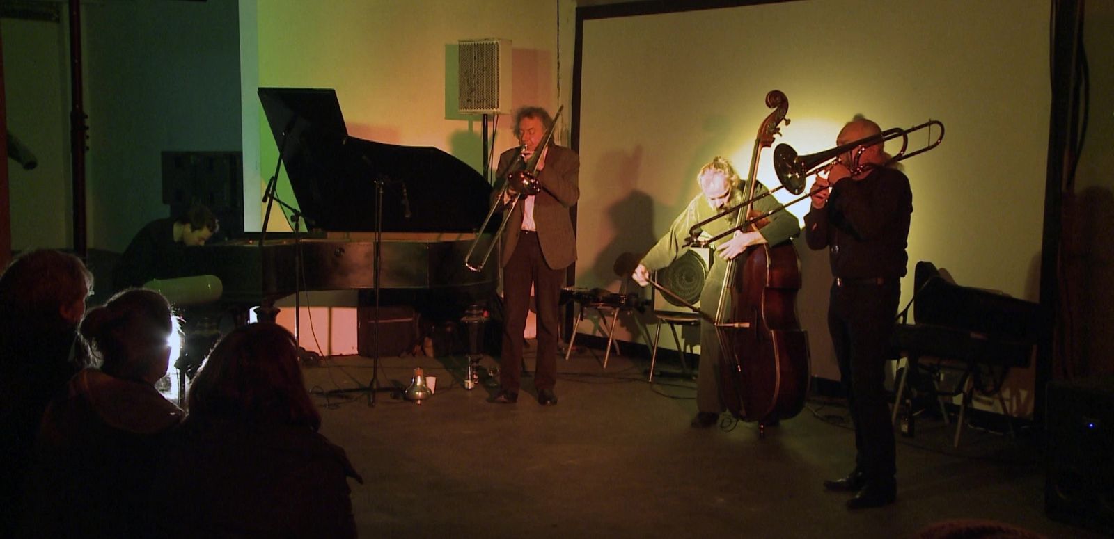 Ein Pianist, zwei Posaunisten und ein Kontrabassist spielen in einem kleinen Raum vor Publikum.