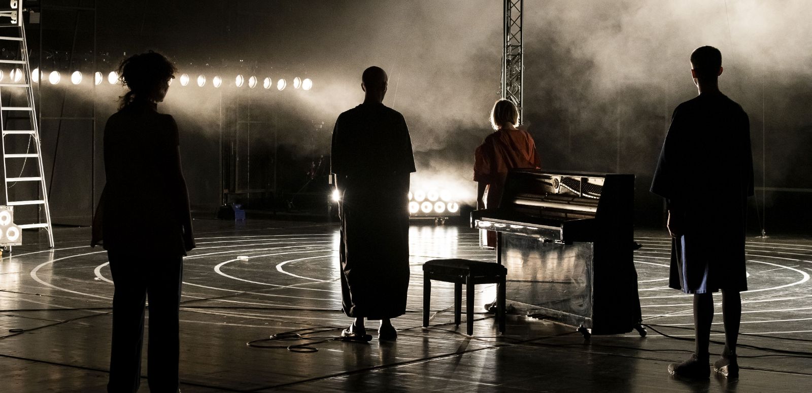 Four people stand backlit on a stage with their backs to the camera, a piano between them.