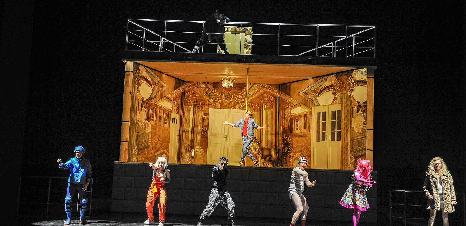 Sechs Personen tanzen vor einem Bühnenbild, das ein golden ausgeleuchtetes, unmöbliertes Zimmer darstellt. In diesem Zimmer tanzt eine weitere Person.