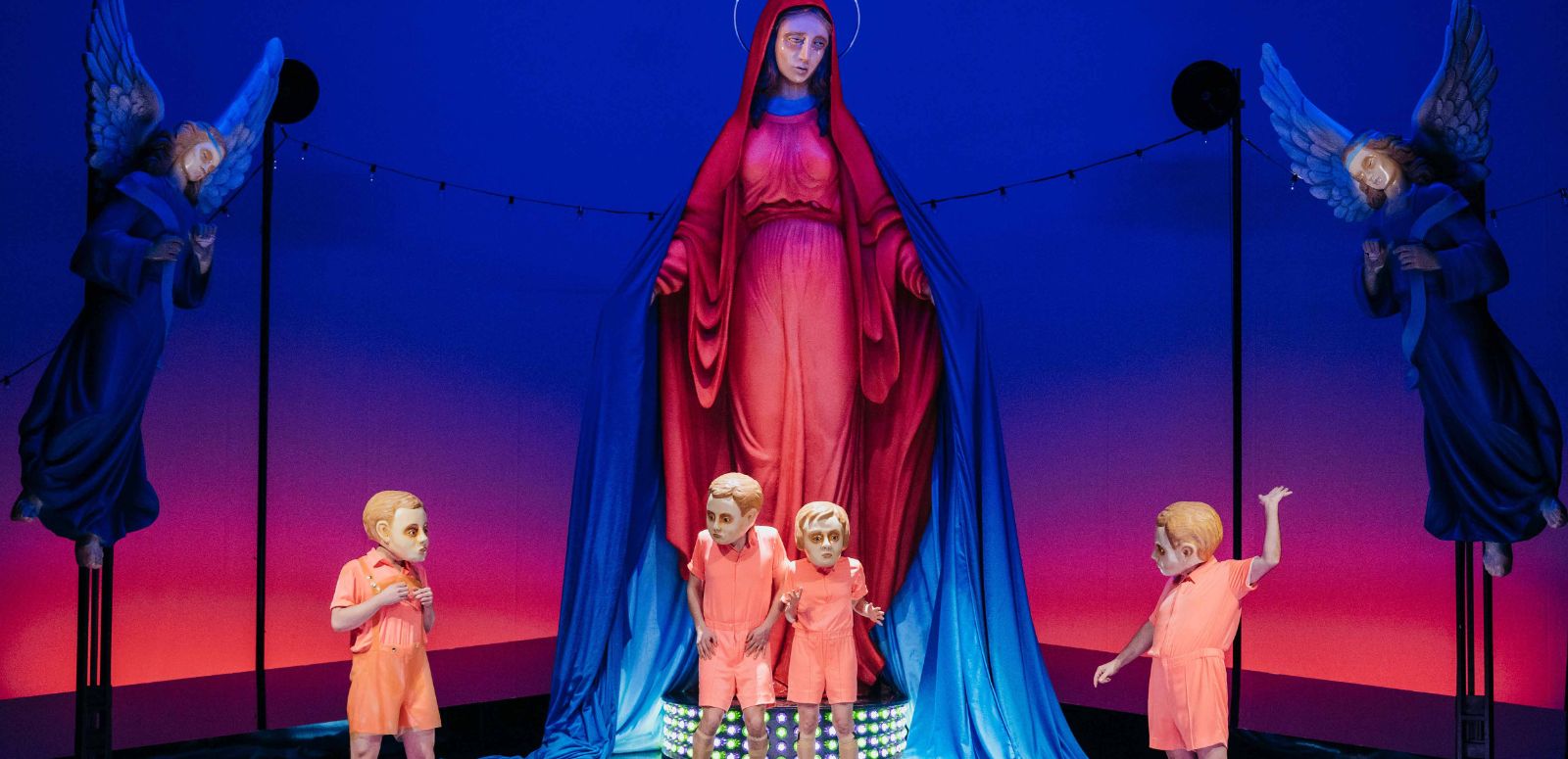Four people in salmon pink costumes and with a mask over their heads stand in front of a huge statue of the Virgin Mary flanked by two angels.