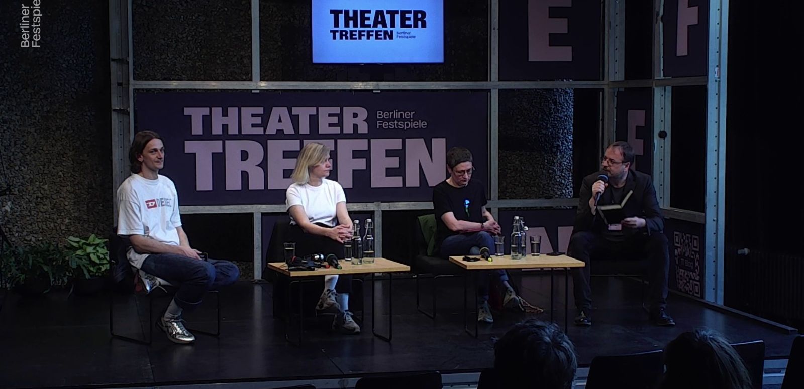 Two women and two men are sitting around a small table, behind them the lettering Theatertreffen. The man on the far right has a microphone in his hand.