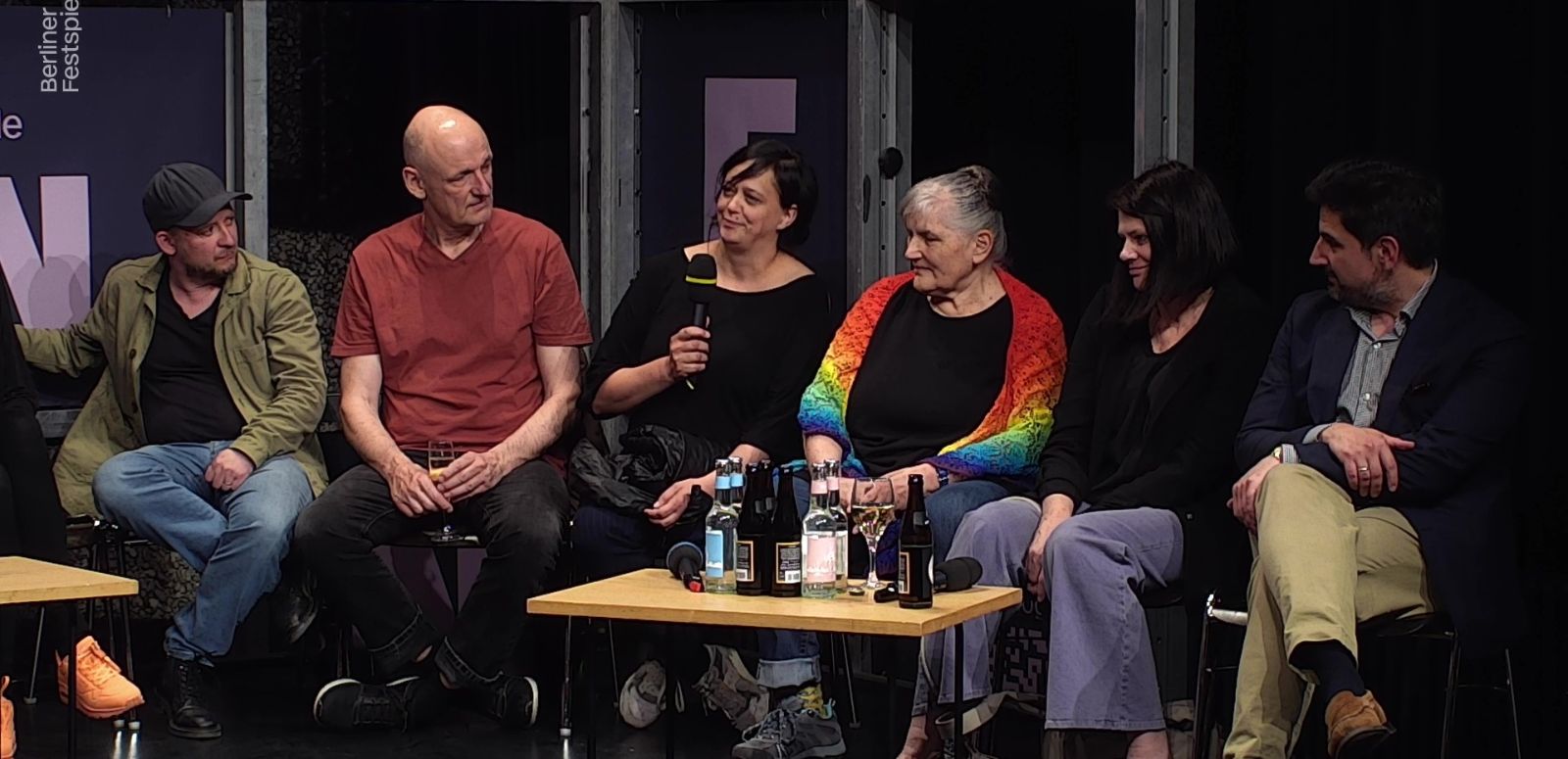 Drei Frauen und drei Männer sitzen um einen kleinen Tisch herum, hinter ihnen der Schriftzug Theatertreffen. Die Frau in der Mitte hat ein Mikrofon in der Hand.
