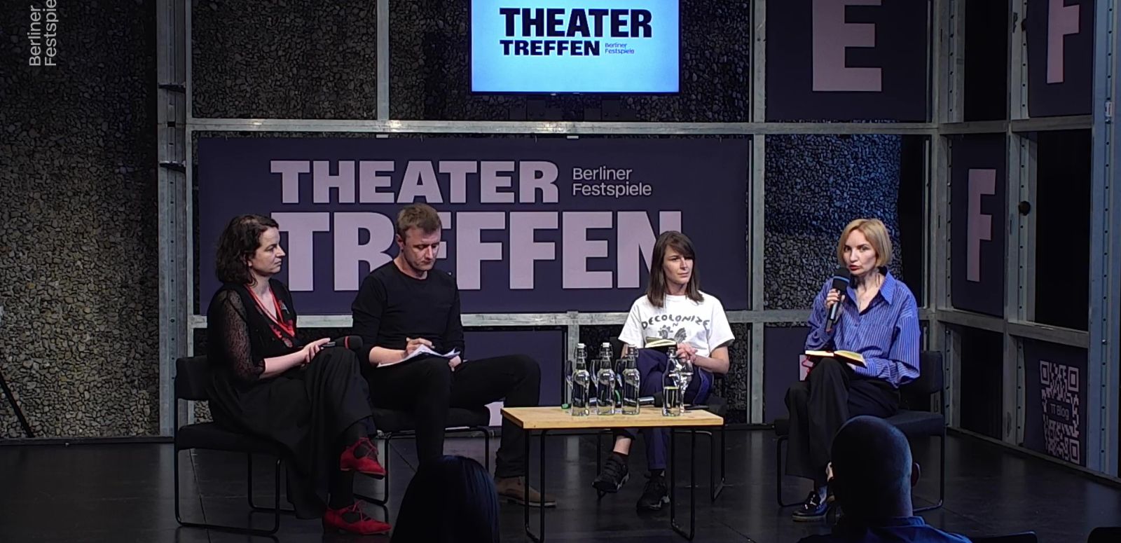 Drei Frauen und ein Mann sitzen um einen kleinen Tisch herum, hinter ihnen der Schriftzug Theatertreffen. Die Frau ganz rechts hat ein Mikrofon in der Hand.