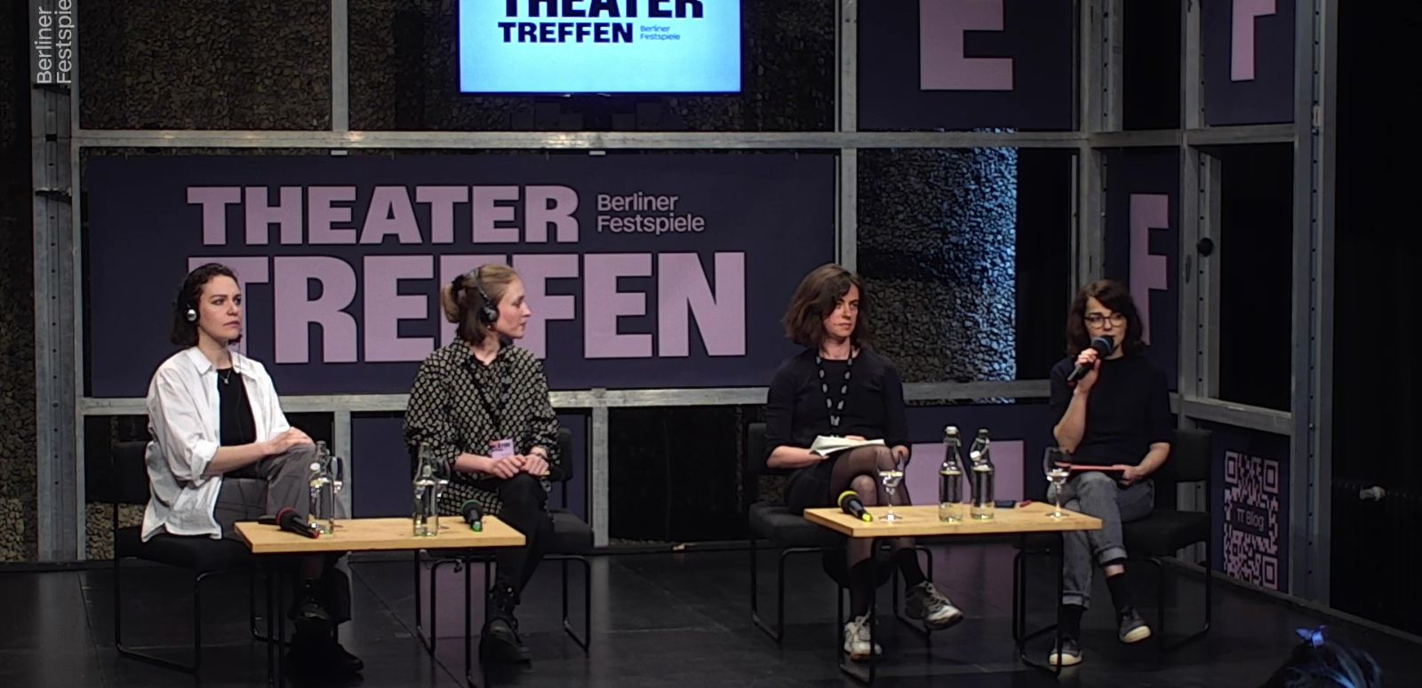 Vier Frauen sitzen an zwei kleinen Tischen, hinter ihnen der Schriftzug Theatertreffen. Die Frau ganz rechts hat ein Mikrofon in der Hand.
