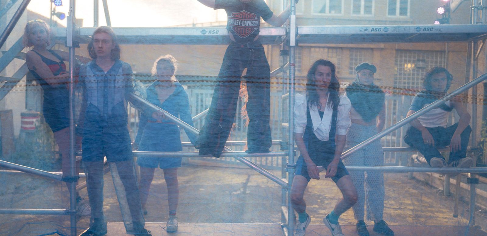 Seven actors  are on a scaffolding with different postures and look through a net.