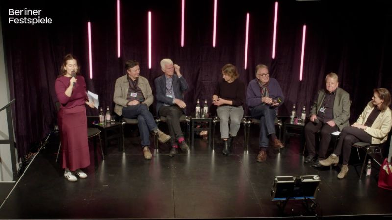 A group of six people sit on a small stage and are introduced by a presenter.