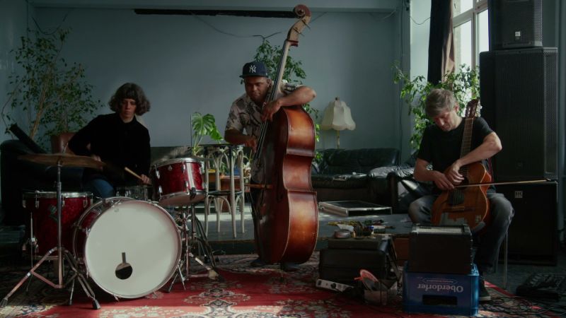 Drei Musiker*innen mit Schlagzeug, Kontrabass und Cello in einem Raum mit Pflanzen, Teppich und Lautsprechern