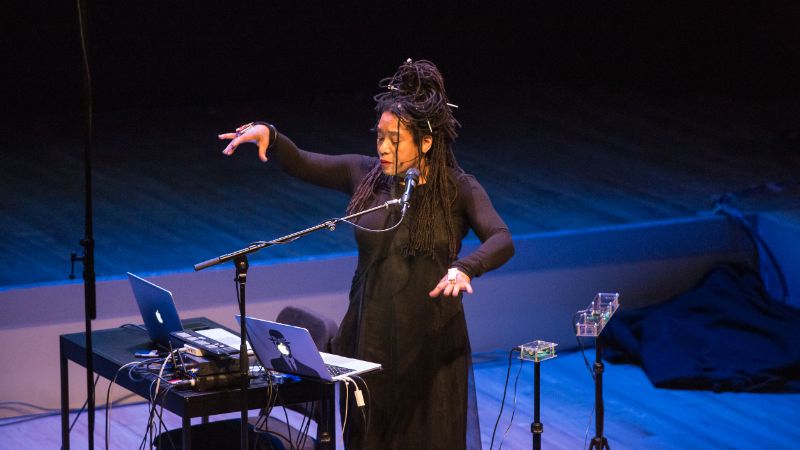Pamela Z uses gestures to control music controllers on a stage.