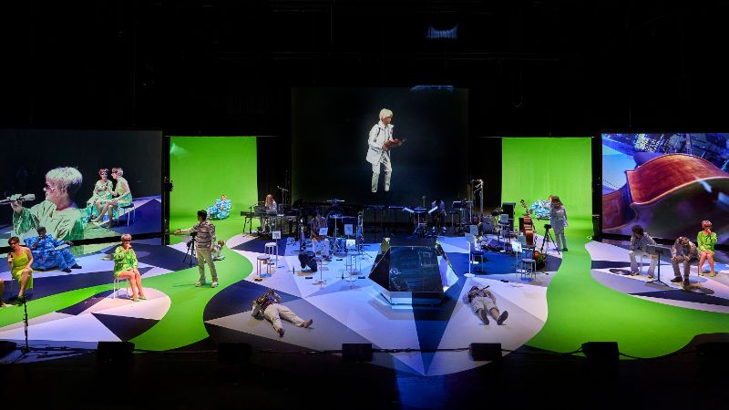Musiker*innen des Ensemble Modern auf der Bühne. Im Hintergrund sind Videoprojektionen von Musiker*innen und Instrumenten.