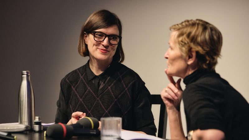 Gisèle Vienne sitzt an einem Tisch und spricht in ein Mikrofon. 