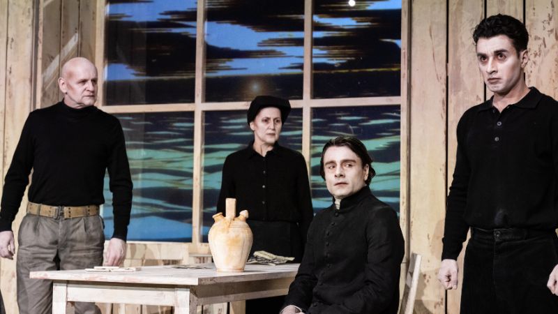 Vier Personen in altertümlich wirkenden schwarzen Klamotten befinden sich in einer Küchentisch-Szenerie, in der alles aus Holz besteht.