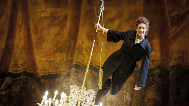 A woman in a blazer‑coat stands smiling on a swinging chandelier
