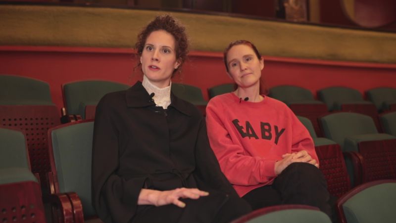 Zwei Personen sitzen nebeneinander in Theatersitzen, eine trägt ein schwarzes Outfit, eine einen roten Pullover mit Schrift.