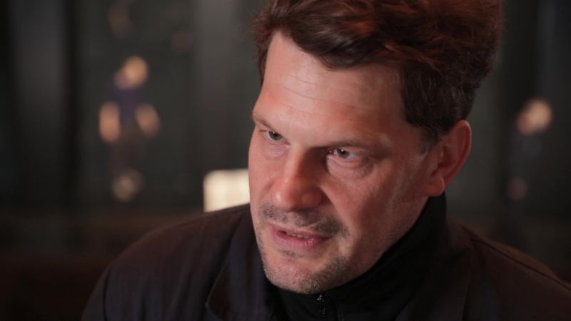 A close-up of a person wearing a dark jacket, seen from the side, with a blurred interior in the background and subdued lighting.