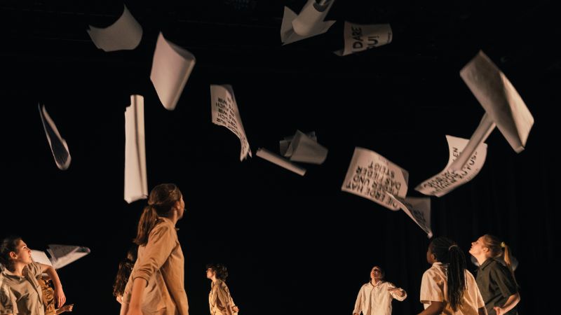 A group of young people, dressed in shades of beige and brown, stand on a stage. Sheets of paper with black lettering fly through the air.
