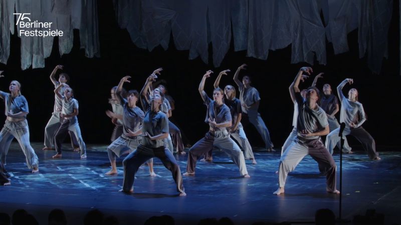 Ensemble von Tanzenden in grauen und weißen Kostümen auf einer Bühne. Die Gruppe führt eine synchronisierte Choreografie mit ausdrucksstarken Armbewegungen unter blauem Licht aus. Über der Bühne hängen stoffartige Elemente. 