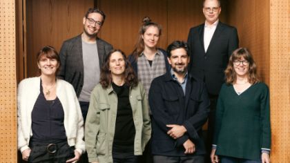 The seven Theatertreffen jury members stand in front of a door of a theatre auditorium and look into the camera.