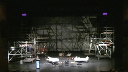 Eva Illouz stands on a stage surrounded by the scaffolding-like set of the play "House".