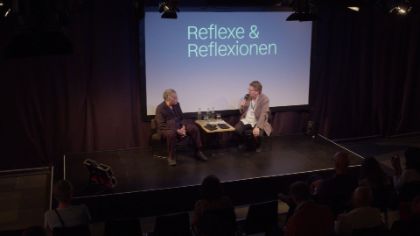 Amos Gitai und Matthias Pees sitzen auf einer Bühne und reden.