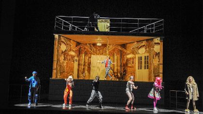 Six people dance in front of a stage set depicting a golden-lit, unfurnished room. Another person dances in this room.
